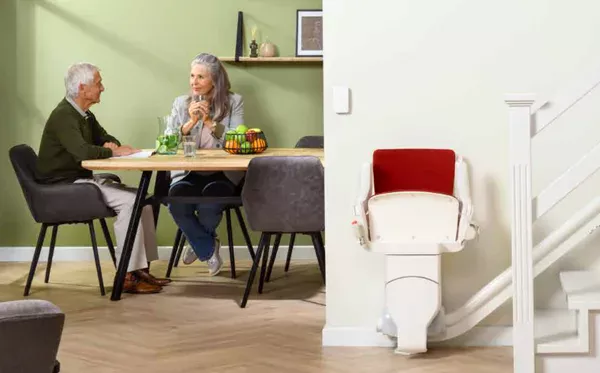 otolift-modul-air Image shows a couple sitting at a table. In the foreground is the Otolift Modul-Air Smart stairlift